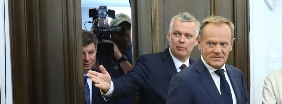 Donald Tusk w garniturze odwraca się w lewo, podczas gdy mężczyzna za nim wskazuje w tę stronę, a w tle widać kamerzystę.