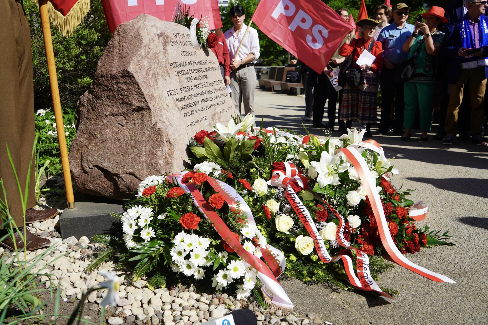 Uroczystość przy pomniku upamiętniającym pierwszą zbrojną demonstrację robotniczą w Polsce z kwiatami i flagami PPS, z udziałem zgromadzonych osób.