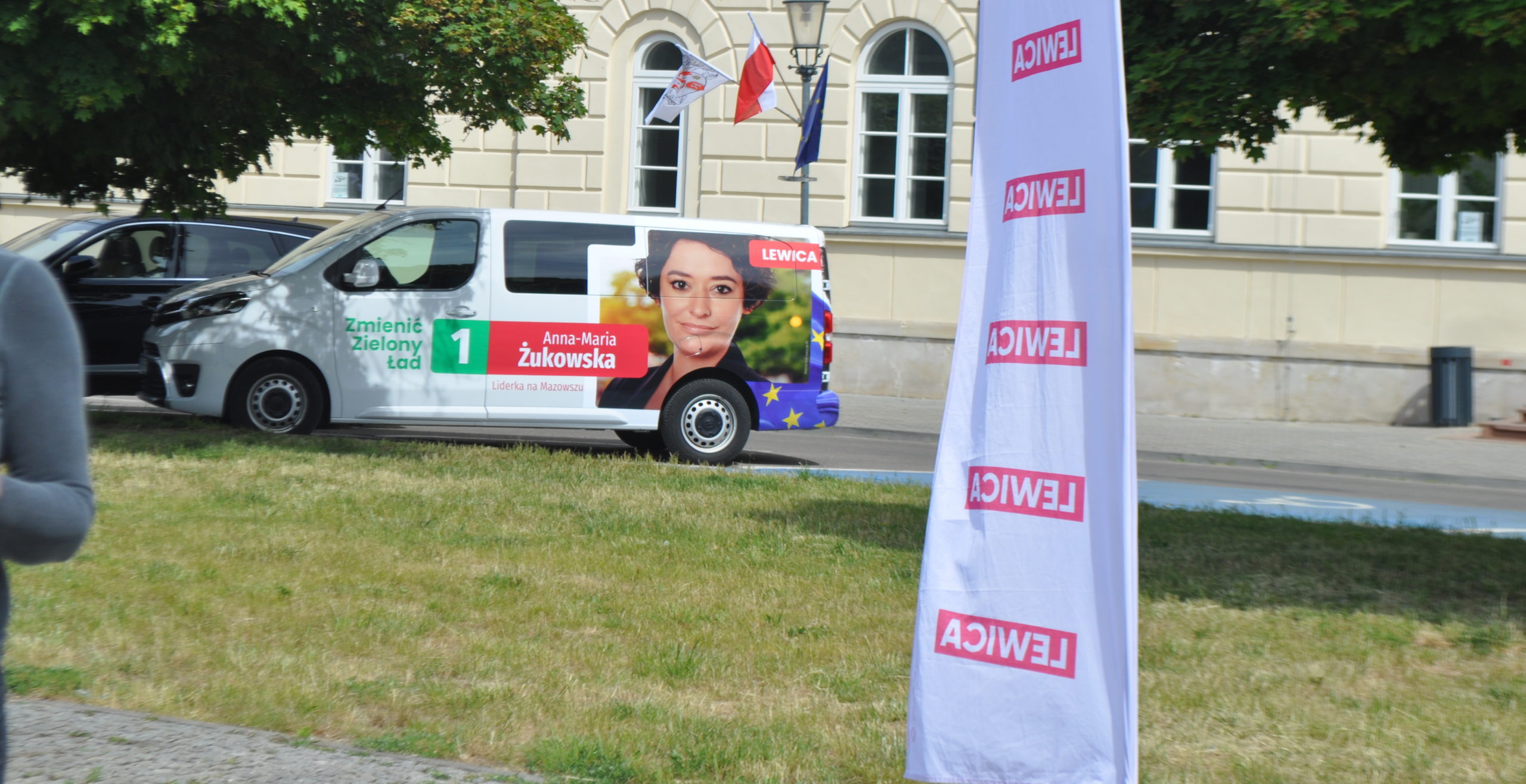 Samochód z reklamą wyborczą Anny-Marii Żukowskiej z partii Lewica zaparkowany przy budynku z flagami.