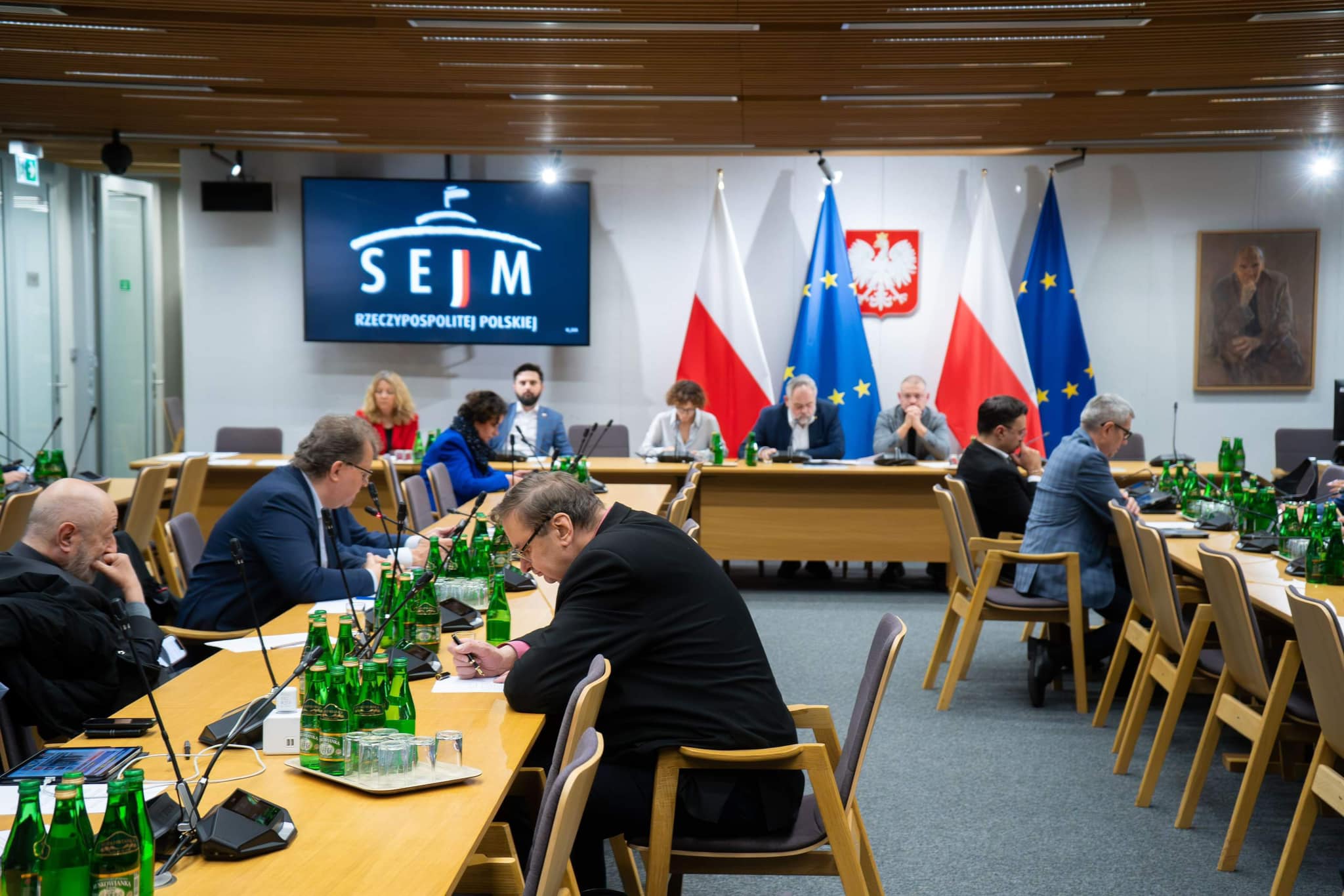 Posiedzenie komisji sejmowej z udziałem parlamentarzystów, na ścianie wyświetlone logo Sejmu oraz widoczne flagi Polski i Unii Europejskiej.