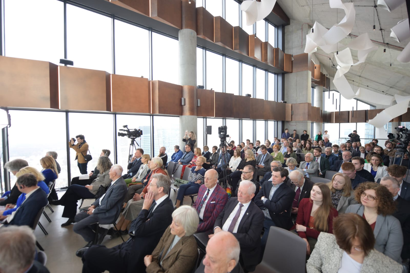 Widok na salę konferencyjną z dużymi oknami, wypełnioną słuchaczami oraz mediami podczas wydarzenia.