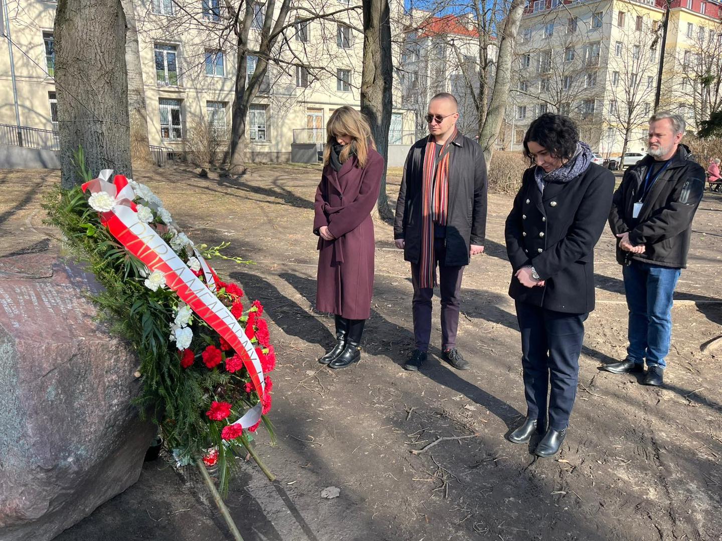 Grupa osób oddaje hołd, stojąc z pochylonymi głowami przy kamieniu upamiętniającym Jolantę Brzeską, obok wieńca z biało-czerwonymi kwiatami.