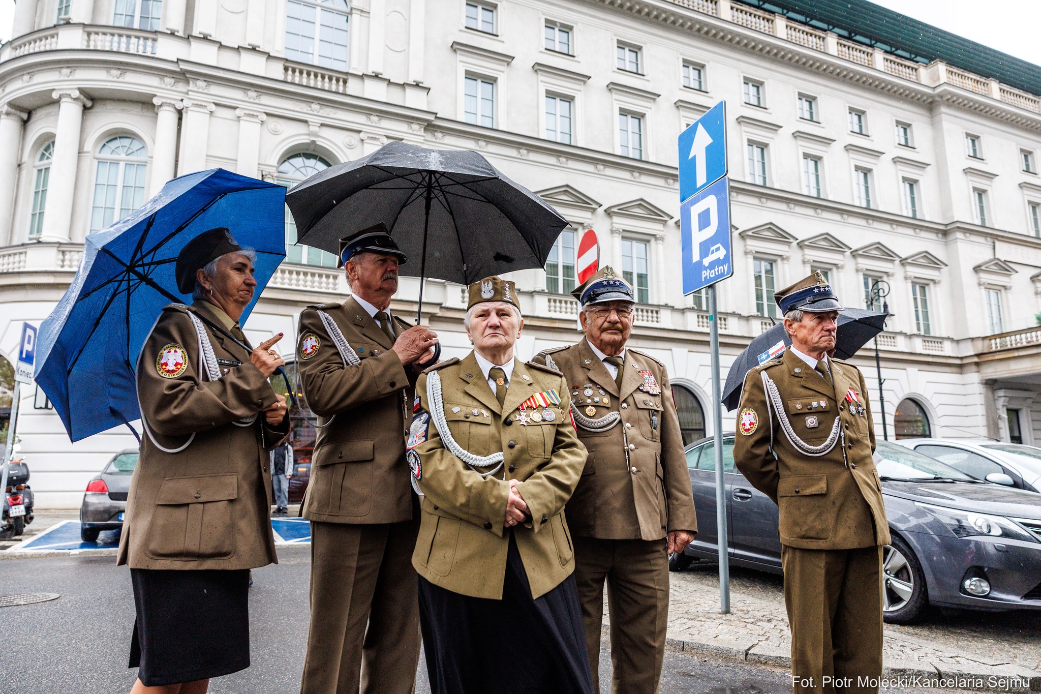 Grupa weteranów w mundurach wojskowych stoi na deszczowej ulicy podczas uroczystości 100. rocznicy Zamachu Majowego.