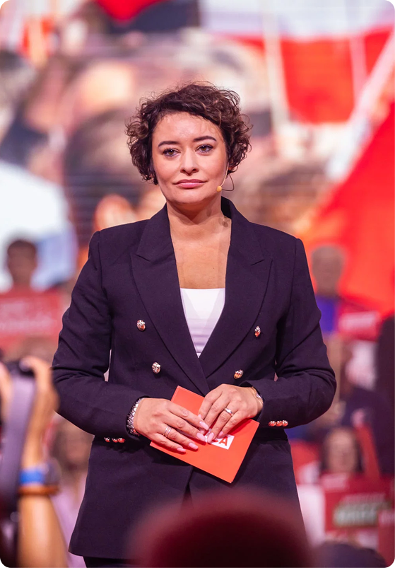 Kobieta w eleganckim garniturze przemawia na tle rozmytego tłumu z czerwonymi transparentami.